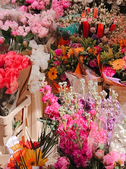 A vibrant display of fresh flowers featuring a variety of blossoms including pink and white roses, bright orange and yellow daisies, and clusters of small pink and purple flowers. The flowers are arra