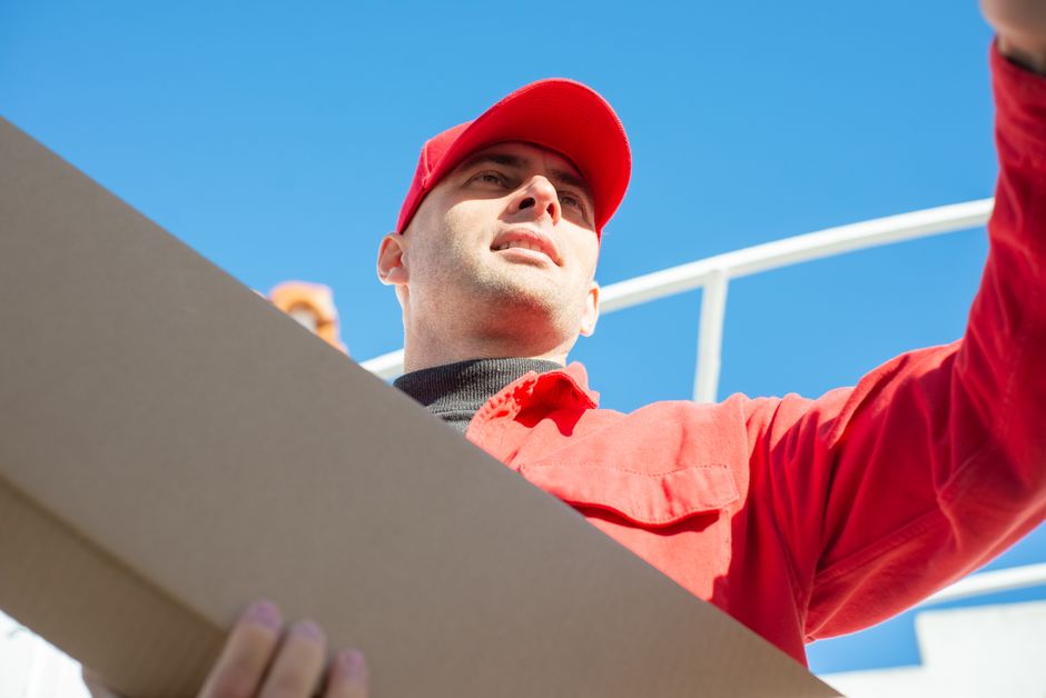 A delivery person wearing a red uniform and cap holding a cardboard box, with a background of clear blue sky and a white railing, indicating outdoor setting. The person's face is partially visible, an
